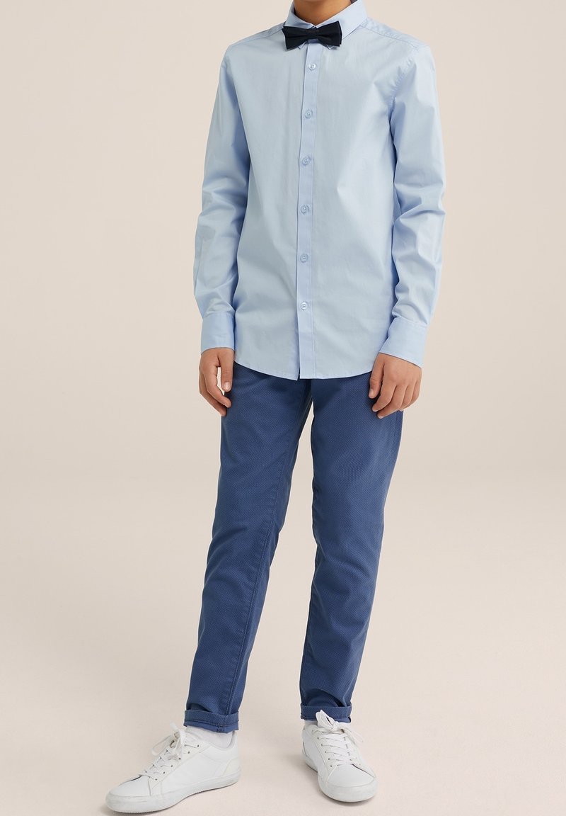 Chemise bleu clair à manches longues et nœud papillon noir, assortie à un pantalon texturé bleu foncé et des baskets blanches.