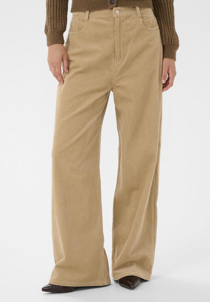 Široke bež kord pantalone u kombinaciji s smešnim pletenim puloverom i tamnim špicastim cipelama, ruke opuštene sa strane.