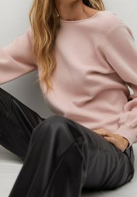 Frau in einem hellrosa Pullover und schwarzen Lederhosen, die mit einem angezogenen Knie vor einem neutralen Hintergrund sitzt.