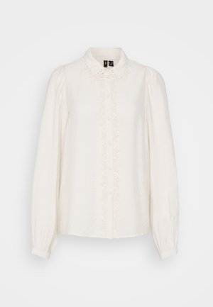 Vit långärmad blus med krage, texturerad tyg, centrala knäppning och blommig broderi framtill.