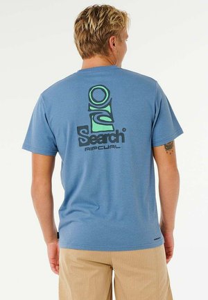 Junger Mann mit kurzen blonden Haaren, der ein blaues Rip Curl Search T-Shirt und beige Hosen trägt, Rückenansicht.