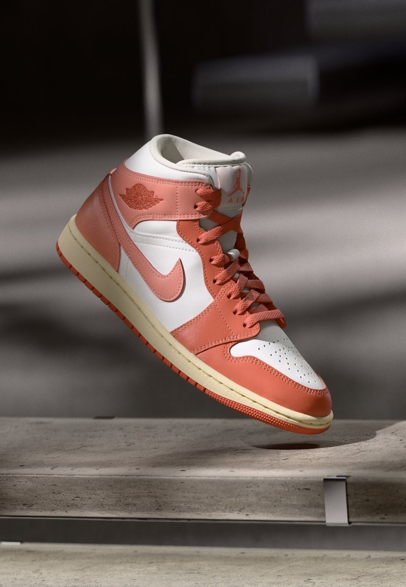 Baskets montantes blanches et corail Air Jordan avec lacets, présentées en lévitation au-dessus d'une surface en béton sur un fond sombre et flou.