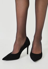 Zapatos de tacón alto de ante negro con punta afilada y un diseño superior abierto, combinados con medias negras transparentes. Textura suave, tacón de aguja delgado.