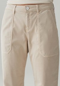 Pantaloni di cotone beige con vita aderente, tasche frontali e chiusura con bottone argentato. Tessuto texturizzato con dettagli di cucitura visibili.