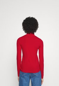Red long-sleeve turtleneck top made of smooth material. The garment features a fitted design and sits at the waist, paired with blue jeans.