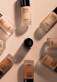 KIKO Unlimited Foundation-flasker arrangert i et sirkulært mønster. Flaskene er laget av frostet glass med svarte lokk; nyansene varierer fra lys til mørk beige.