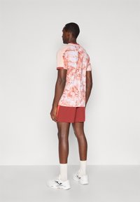 Σετ ανδρικών αθλητικών ρούχων: μπλούζα κοντομάνικη σε ροζ χρώμα με αφηρημένο κόκκινο σχέδιο, συνδυασμένη με κόκκινες βερμούδες. Τα λευκά αθλητικά παπούτσια και οι γκρι κάλτσες ολοκληρώνουν το σύνολο.