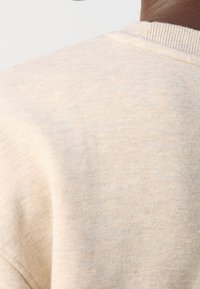 Sudadera de color beige claro con una tela de algodón texturizada, que presenta un cuello redondo y un escote de canalé. Las costuras visibles añaden un detalle sutil.