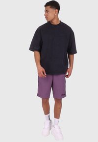 Oversize czarny bawełniany t-shirt, fioletowe szorty z bocznymi paskami oraz białe sneakersy. Swobodny wygląd z minimalną marką i luźnym krojem.