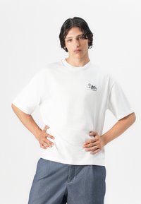 White cotton t-shirt with short sleeves, featuring a small black mountain and sun graphic on the left chest. Loose fit and round neck.