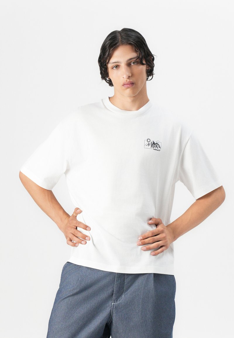 T-shirt en coton blanc à manches courtes, avec un petit graphique de montagne noire et de soleil sur la poitrine gauche. Coupe ample et col rond.