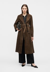 Femme portant un long manteau en suède marron ceinturé, un pantalon large noir, des ballerines pointues noires, et tenant une pochette noire matelassée.