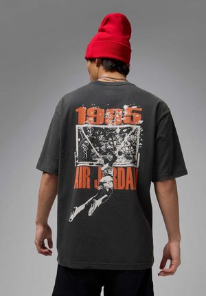 Dark gray oversized t-shirt with a basketball graphic on the back. Features bold orange text "1985" and "AIR JORDAN" in stylized font.