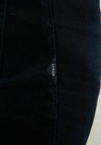 Pantalones de denim oscuro con una textura suave, que cuentan con una etiqueta sutil en el costado etiquetada como "CHASIN". El diseño es ajustado, destacando una silueta estilizada.