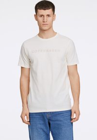 Valkoinen t-paita pyöreällä kaula-aukolla, jossa on kohotettu teksti "COPENHAGEN" rinnassa. Yhdistetty sinisiin farkkuihin.