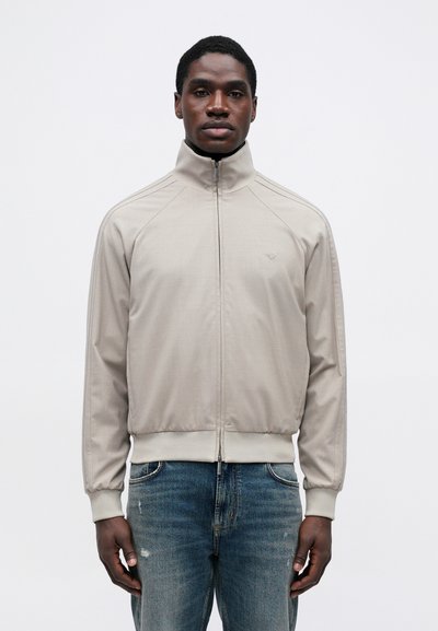 Ανοιχτό γκρι ζακέτα με φερμουάρ, σταντ γιακά, ριμπ μανσέτες και εφαρμοστή μέση. Διαθέτει διακριτές ραφές στους ώμους και ένα μικρό λογότυπο.