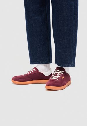 Persoon die donkerblauwe spijkerbroek, witte sokken en bordeauxrode sneakers met oranje zolen draagt, staand op een lichtgrijze vloer.