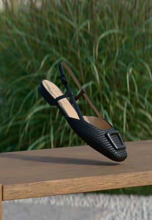 Μαύρο πλεκτό φλατ παπούτσι με λουρί στο πίσω μέρος και μεγάλο ορθογώνιο αγκράφα, σε ξύλινη επιφάνεια, φόντο με πράσινο γρασίδι.