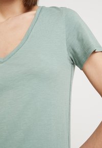 Kortärmad V-ringad skjorta i ljusgrön tyg med en jämn textur och subtila sömdetaljer längs halslinjen och sidorna.