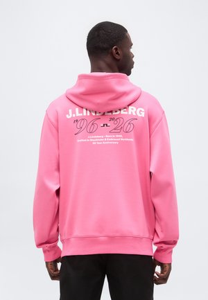 Mann in einem pinkfarbenen Hoodie mit dem Text J.LINDEBERG 1996-2026 und Jubiläumsdetails auf dem Rücken, trägt schwarze Hose.