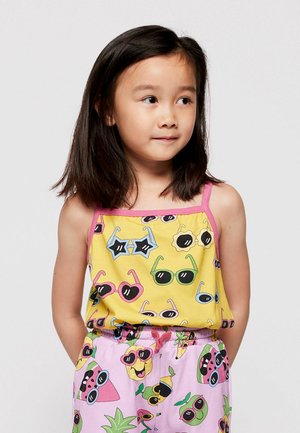 Jeune fille aux cheveux noirs et raides, portant un haut jaune et un short rose décoré de lunettes de soleil colorées et de personnages fruités souriants.