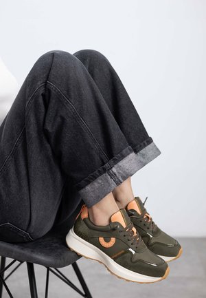 Zapatilla verde con acentos naranjas, lados texturizados y suela blanca gruesa; combinada con pantalones de mezclilla oscuros con dobladillo.