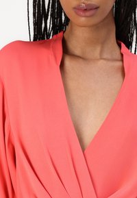 Blusa avvolgente corallo con scollo a V profondo, caratterizzata da maniche fluide e una texture liscia. Il tessuto ha una leggera lucentezza e un leggero drappeggio.