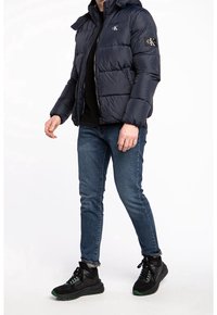 Mörkblå puffjacka med huva, med horisontella quiltade sektioner och en liten logotyp på bröstet. Matcher med mörka jeans och svarta sneakers.