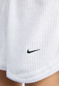 Detailní záběr bílých sportovních šortek s černým logem Nike na dolní straně, zdůrazňující texturovanou tkaninu a lemování.