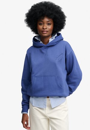 Vrouw met afro-kapsel die een blauw capuchontrui draagt over een blauw gestreept overhemd en witte broek, staand voor een effen achtergrond.