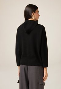 Czarny dziany sweter z kapturem, z prążkowanymi mankietami i obszyciem. Prosty, przytulny design o luźnym kroju, zestawiony z szarymi spodniami.