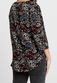 En kvinna i en svart blommig blus med färgglada mönster står på sidan och visar baksidan av skjortan och mörka jeans.