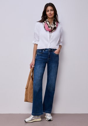 Femme portant une chemise blanche à boutons, un jean bleu à jambes larges, une écharpe colorée, des baskets argentées, tenant un sac fourre-tout en daim beige, debout contre un mur uni.