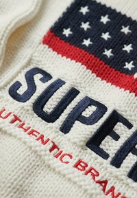 Maglione lavorato a maglia con una base bianco sporco, ricamo blu con la scritta "SUPER" e un motivo con bandiera rossa, bianca e blu decorata con stelle.