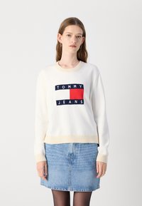 Hvit strikket genser med beige detaljer som har en marineblå og rød logo-patch som leser "TOMMY JEANS." Kombinert med en denimskjørt.