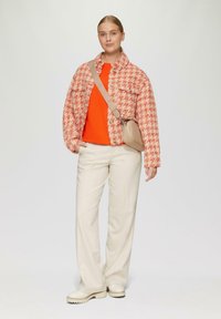 Rot-weiße Hahnentrittmuster-Jacke mit zwei Brusttaschen, kombiniert mit einem orangefarbenen Rippenoberteil, beigen Hosen und cremefarbenen klobigen Schuhen.