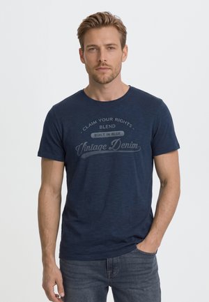 Un homme porte un tee-shirt bleu marine à motif graphique et un jean foncé sur un fond clair uni, une main dans la poche.