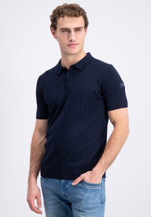 Jeune homme aux cheveux bouclés portant un polo côtelé bleu marine à manches courtes et un jean bleu clair, debout avec une main dans la poche.