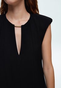 Top de tela negra con escote en V y un acento dorado cerca del cuello. Mangas cortas y estructuradas con sutiles pliegues en el frente.