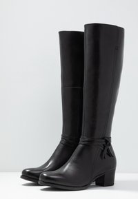 Botas de cuero negra hasta la rodilla con una textura suave, punta redondeada, tacón cuadrado y un adorno de lazo decorativo en el tobillo.