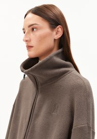 Femme aux cheveux bruns raides portant une veste en polaire taupe à col montant, regardant à gauche sur un fond blanc uni.
