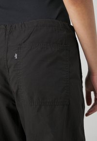 Personne portant un pantalon noir avec une poche arrière et une étiquette avec le logo Levi's visible, debout avec la main droite détendue le long du corps.