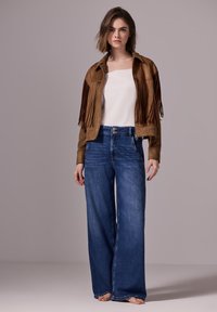 Brun mockajacka med fransdetaljer, vit topp och vida blå jeans med hög midja och knappstängning. Svala texturer och avslappnad stil.