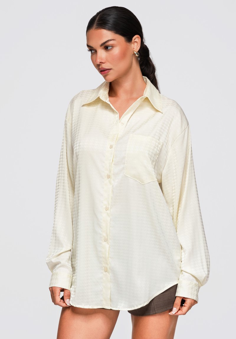 Femme aux cheveux foncés en queue de cheval basse, portant une chemise oversize crème à manches longues boutonnée avec une poche poitrine.