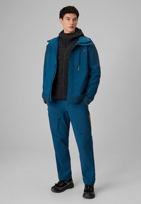 Jeune homme portant une veste et un pantalon d'extérieur bleus avec une veste intérieure noire et des chaussures de randonnée noires, debout les mains dans les poches.