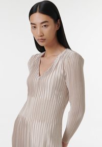 Robe plissée rose clair avec des manches longues et un décolleté en V. Fabriquée dans un tissu lisse et brillant avec une texture verticale sur l'ensemble.