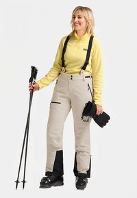 Beige waterdichte skibroek met zwarte accenten, verstelbare bretels, zijritsen en een hoge tailleband, gecombineerd met een geel fleece shirt.