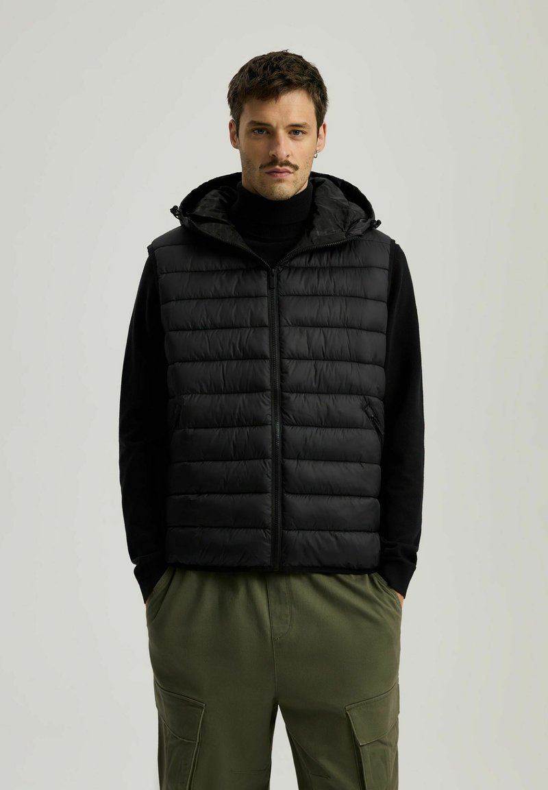 Czarna kamizelka puchowa z zamkiem z przodu i kapturem, o stebnowanej teksturze i bocznymi kieszeniami. Nosi się ją na czarnym golfie i zielonych spodniach.