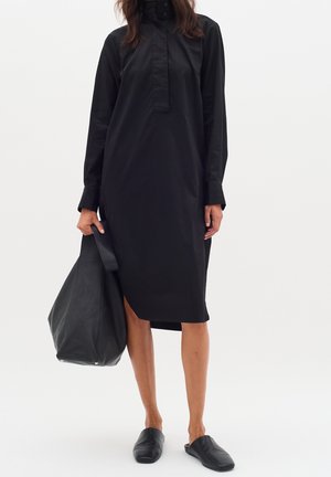 Kobieta w długiej czarnej sukience koszulowej z długimi rękawami, trzyma dużą czarną torbę typu tote i ma na stopach czarne buty wsuwane na płaskim tle.