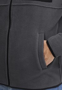Chaqueta de felpa gris con cremallera, que presenta un acabado texturizado, bolsillos laterales y detalles en negro en el collar y los puños.